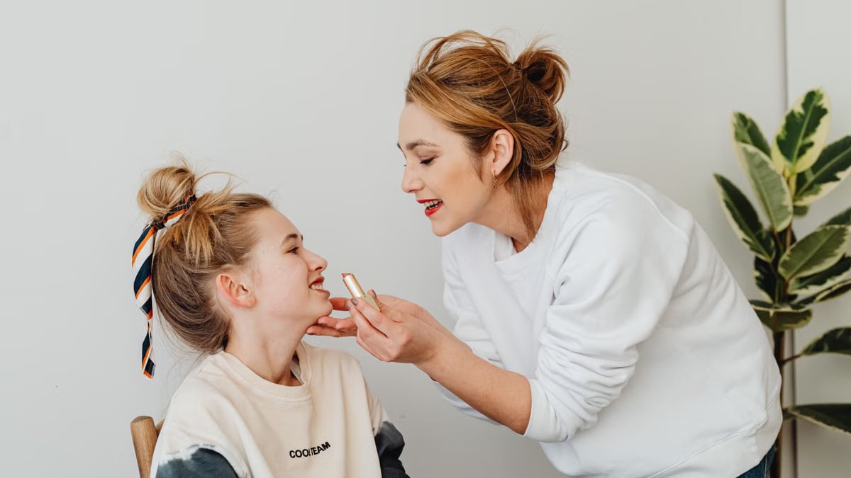 A mother lovingly applies lipstick to her smiling daughter indoors, capturing a tender family bonding moment.